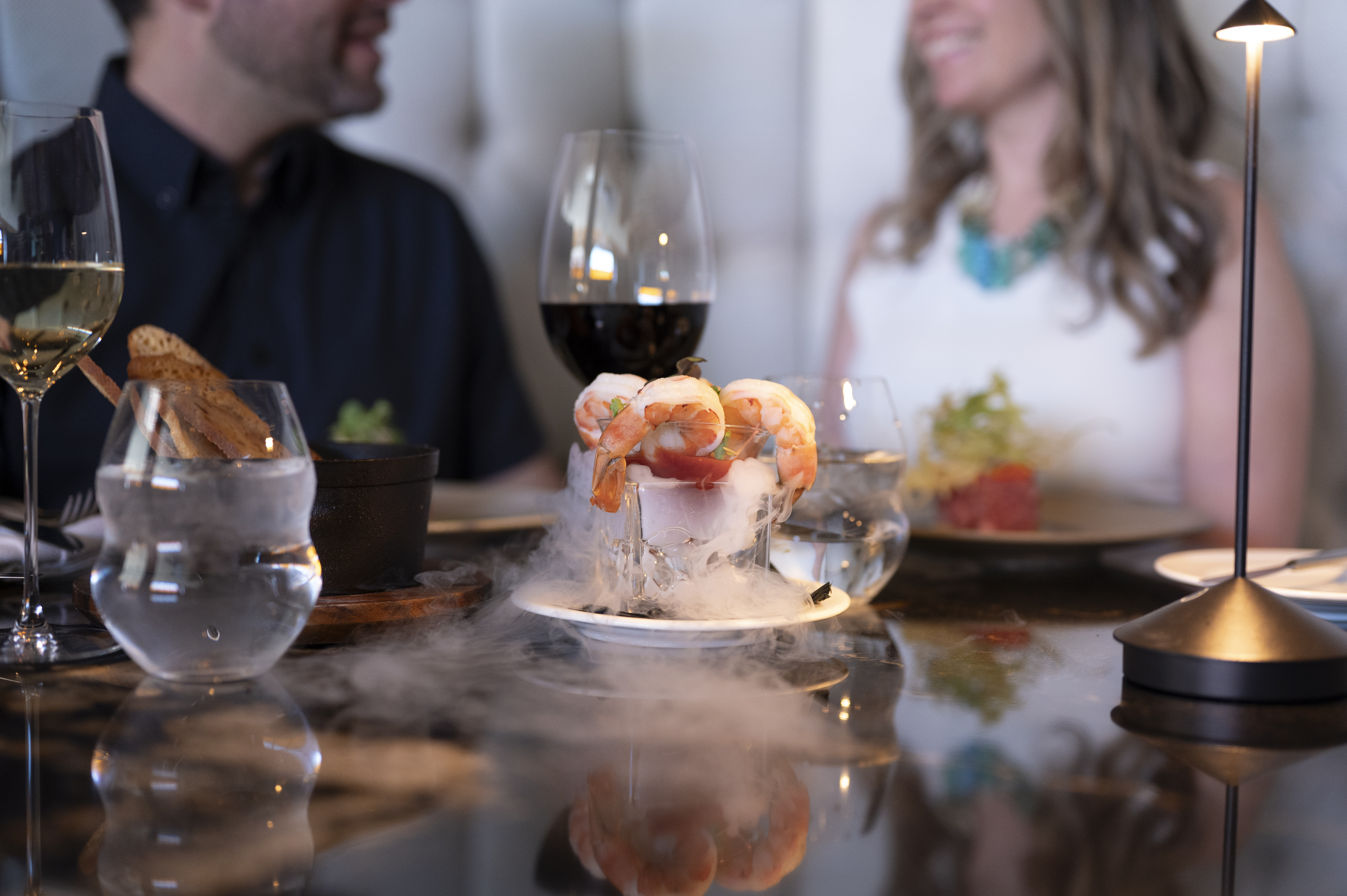 A close-up of a shrimp cocktail amidst smoke and couples dining in the background
