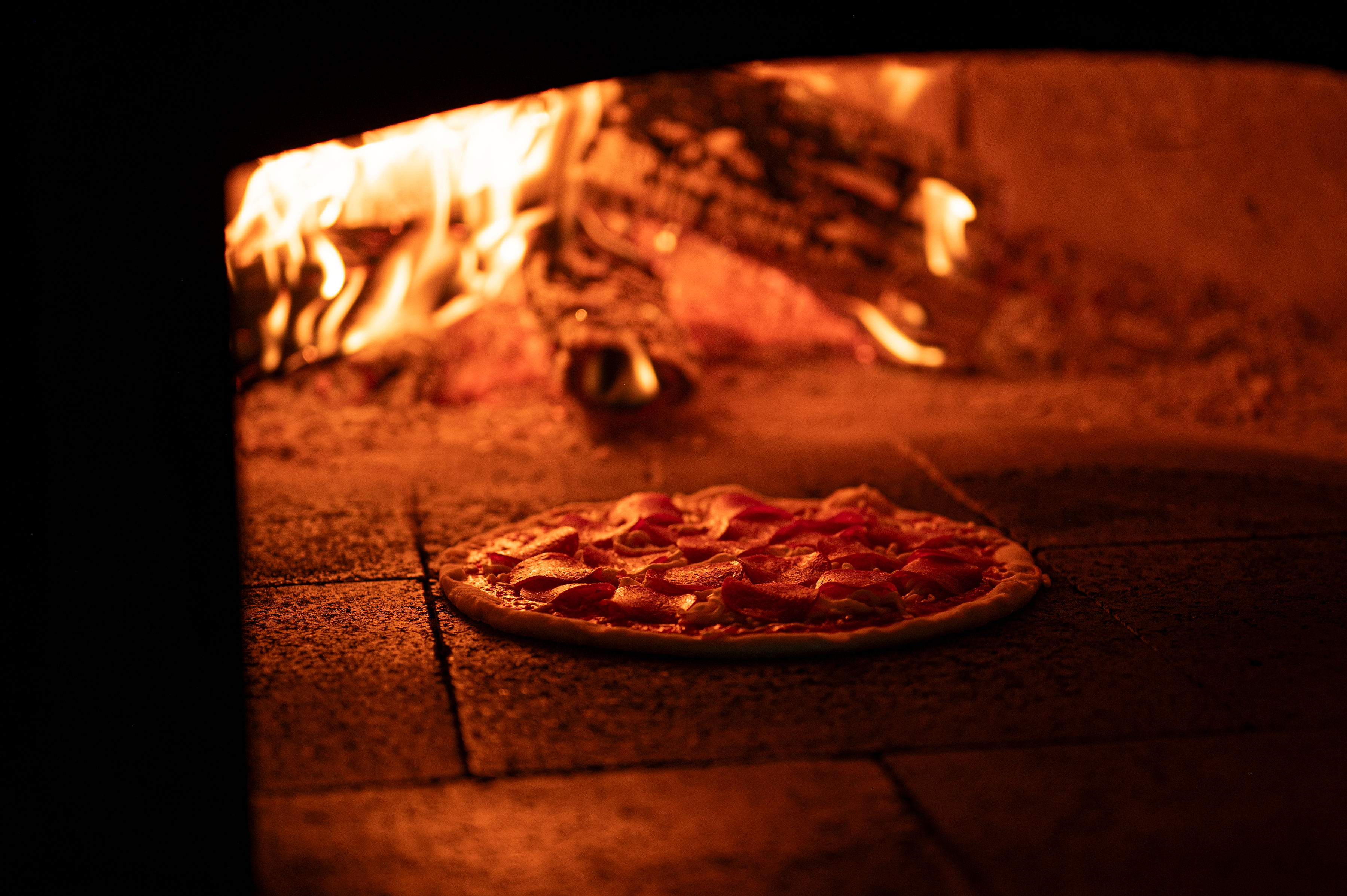 Uncooked pepperoni pizza sits inside a wood-fired oven glowing with flames