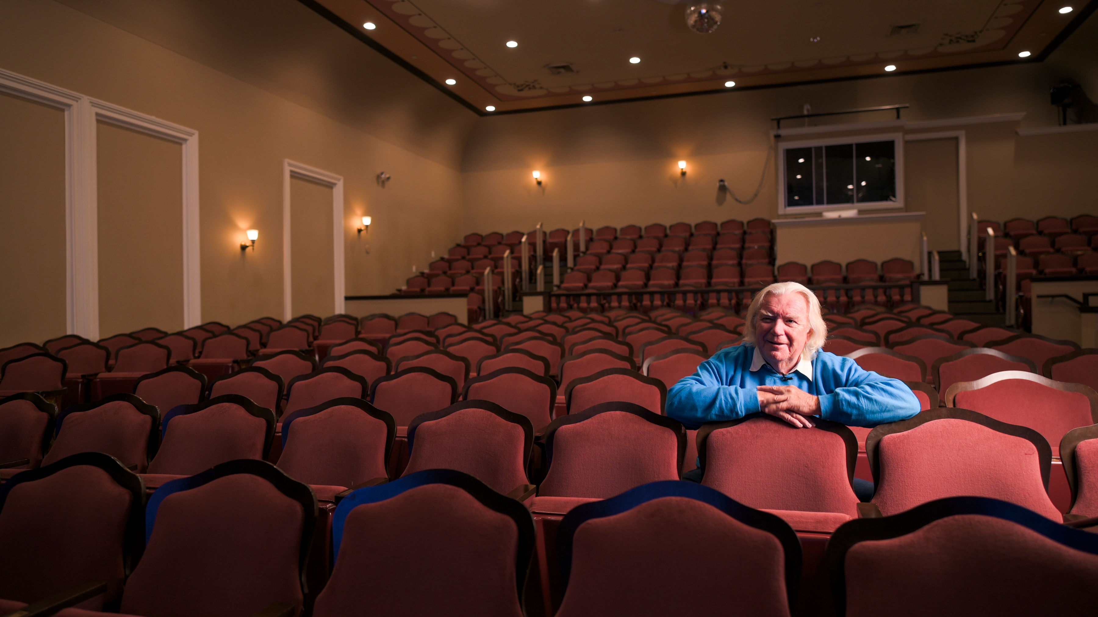 Man with white hair in a blue shirt sits in a red velvet seat in a theater