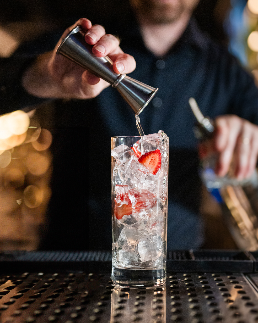 Bartender pours liquid from a jigger into a tall glass with ice and strawberries