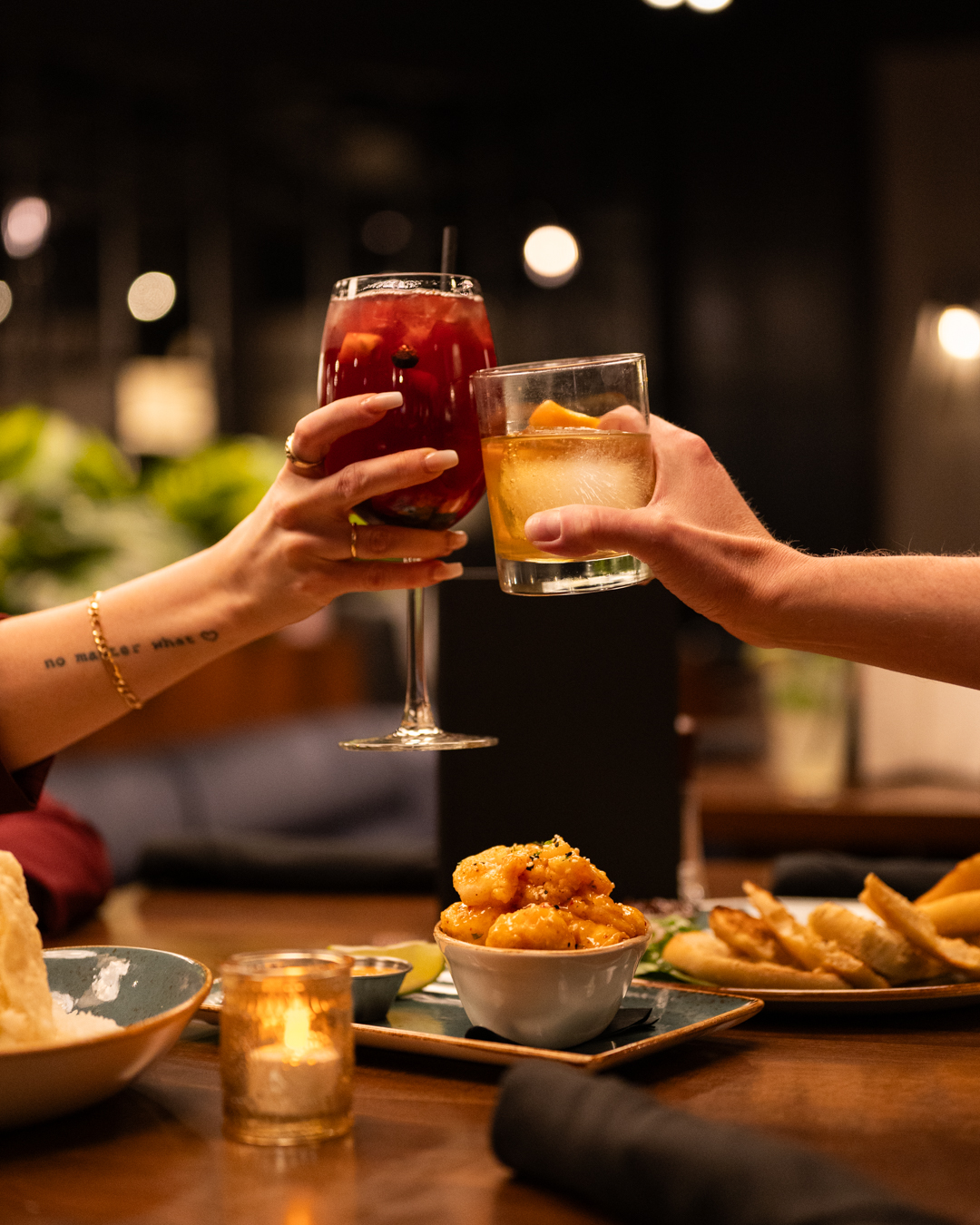 Two people clinking glasses, one red cocktail, one amber cocktail over food