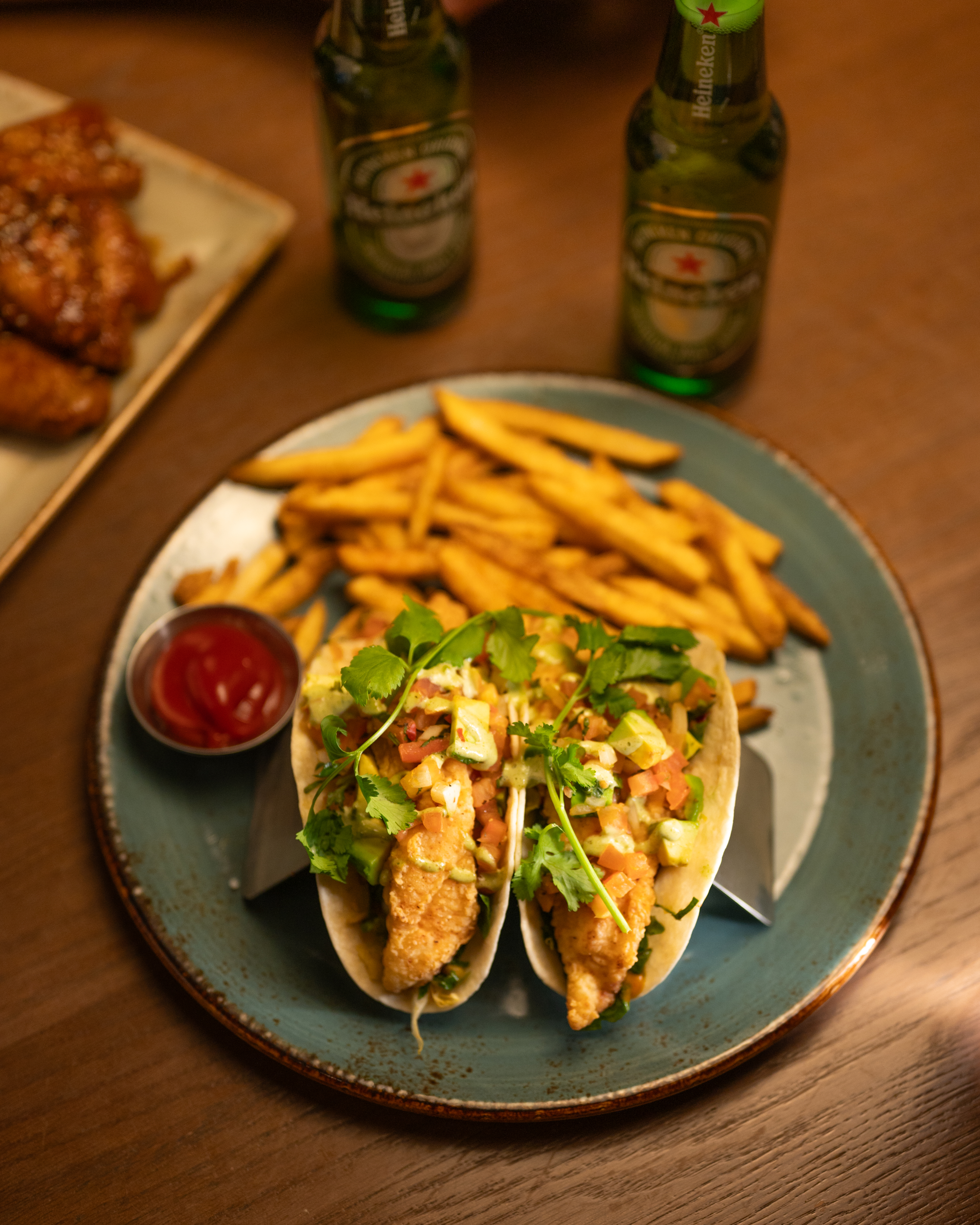 Two Milestones fish tacos with fries, ketchup, and two bottles of beer on a wooden table