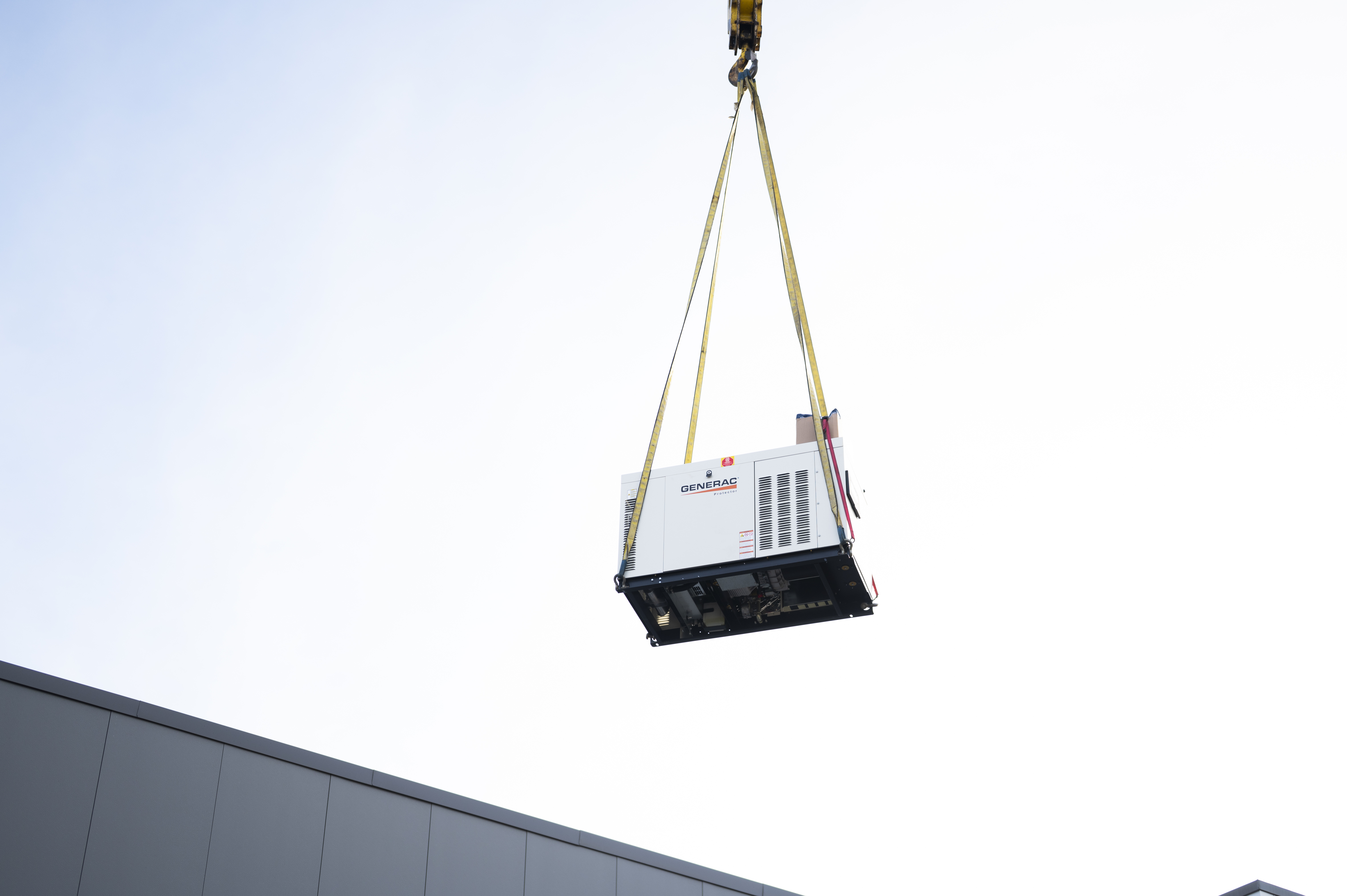 Heavy Generac generator lifted by crane against a clear sky above a building