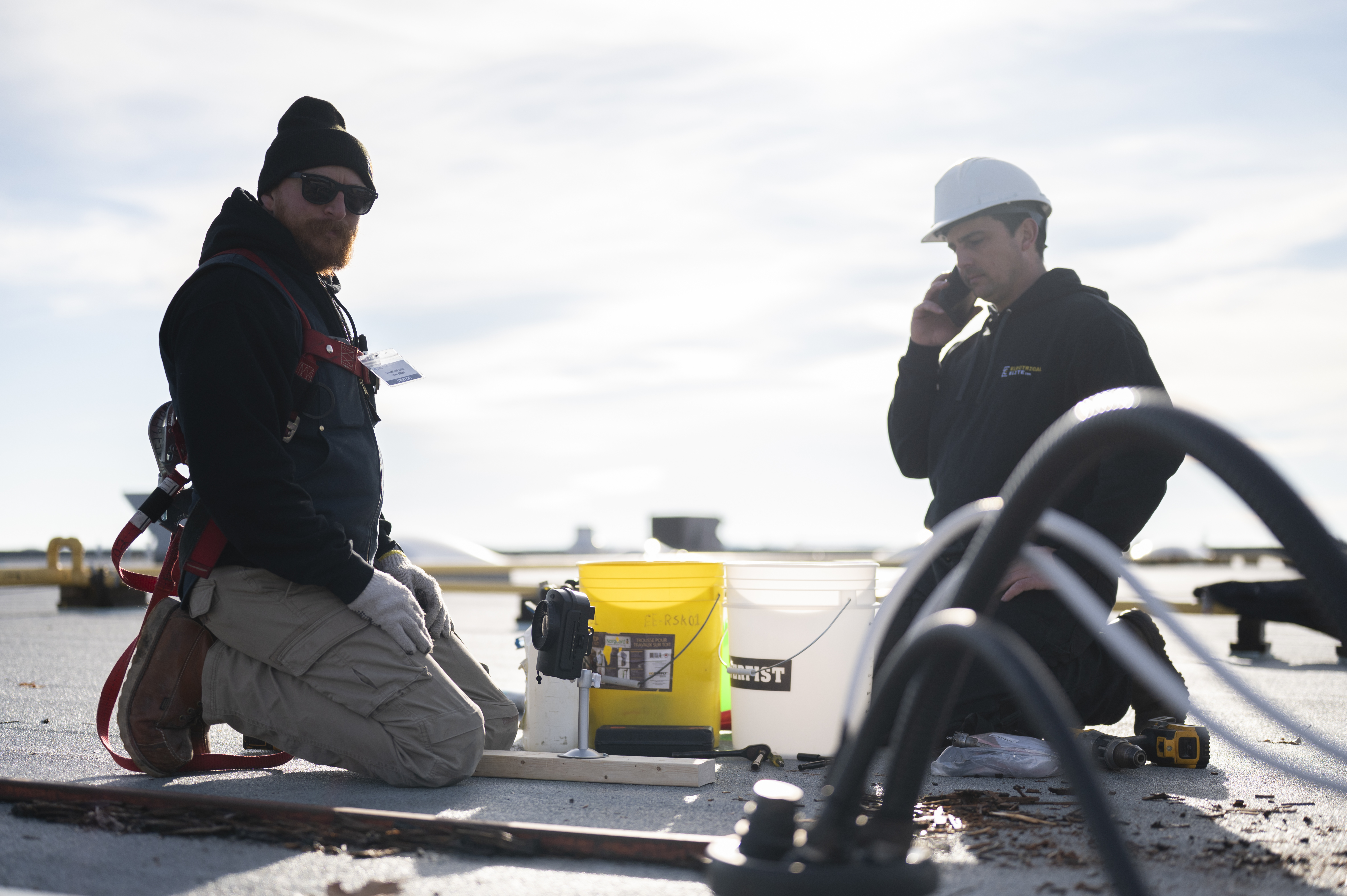 Two construction workers on a rooftop, one on phone, surrounded by tools