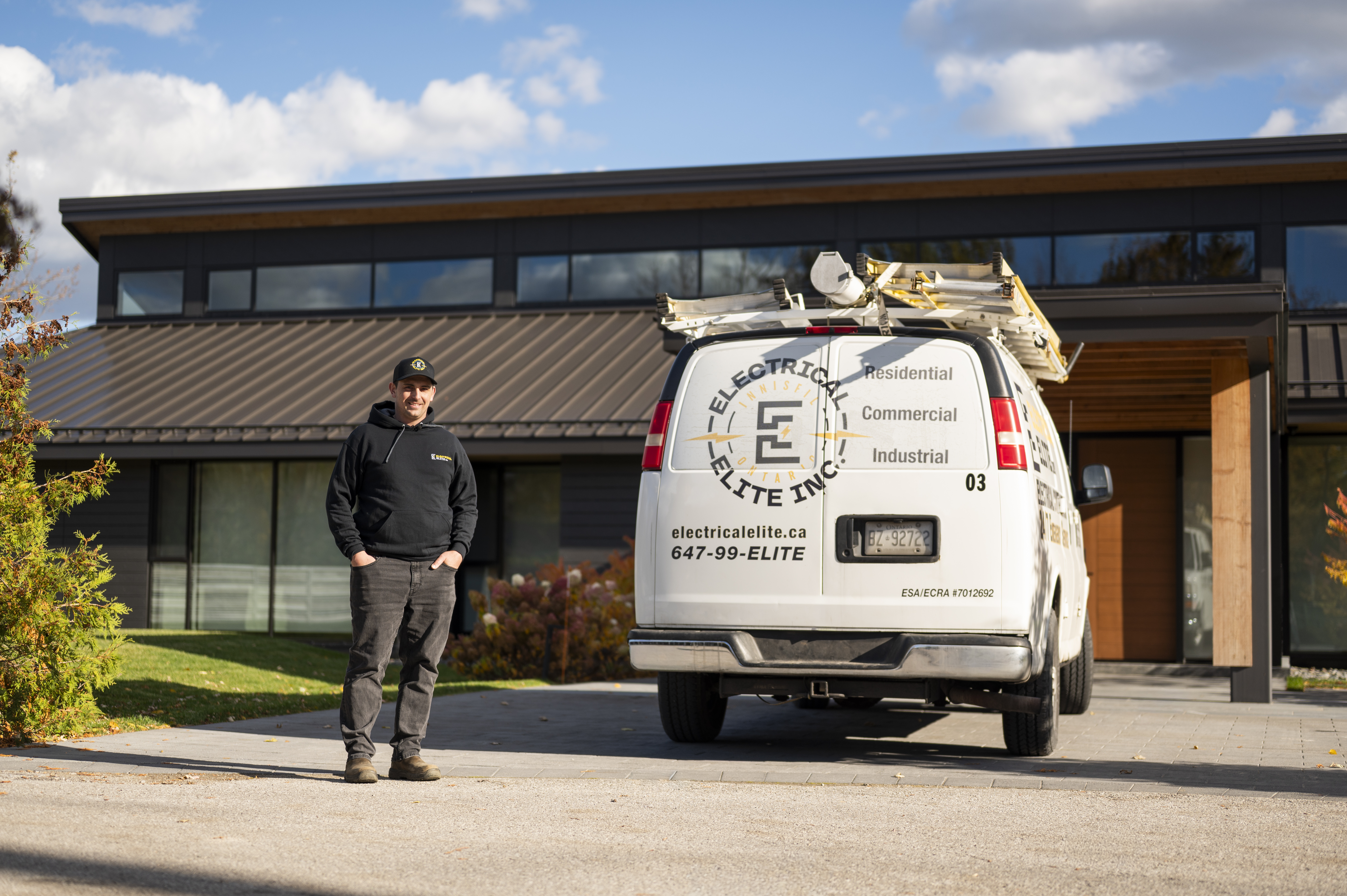 Electrical Elite technician in black hoodie standing beside a company van outside a modern building