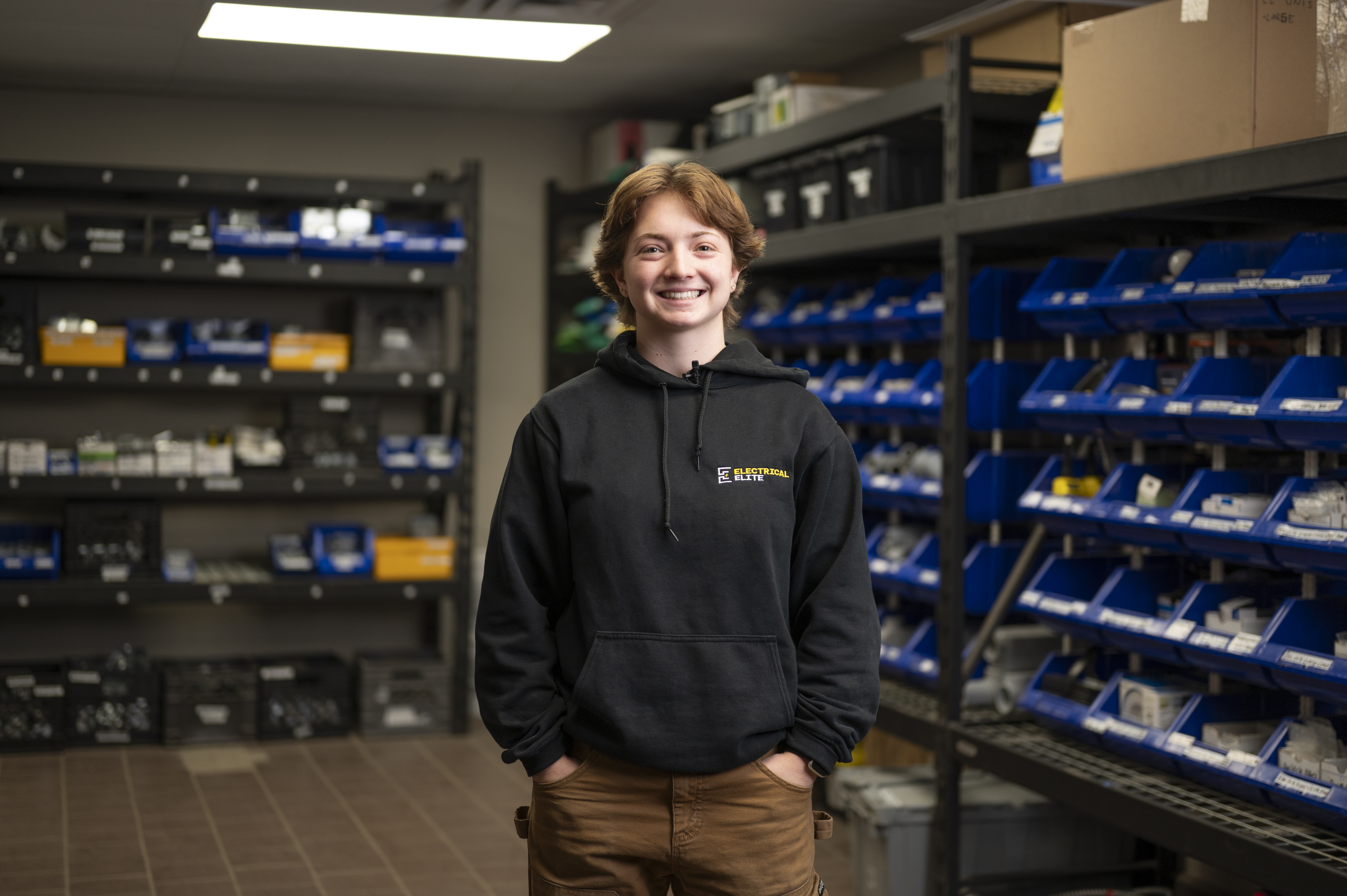 Smiling young person wearing an Electrical Elite hoodie in a parts warehouse