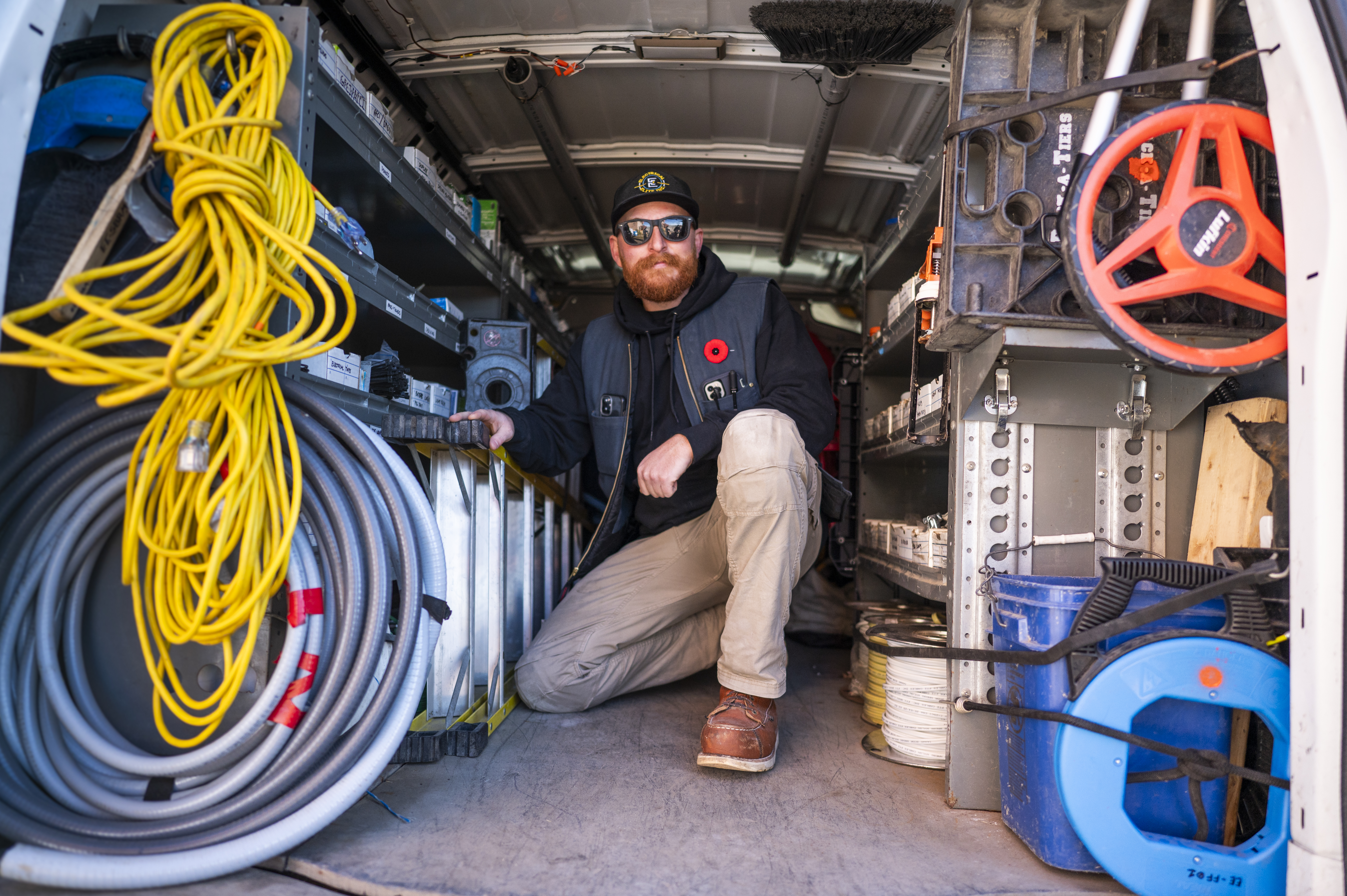 Male electrician with beard, sunglasses, and work vest kneeling inside a packed service van