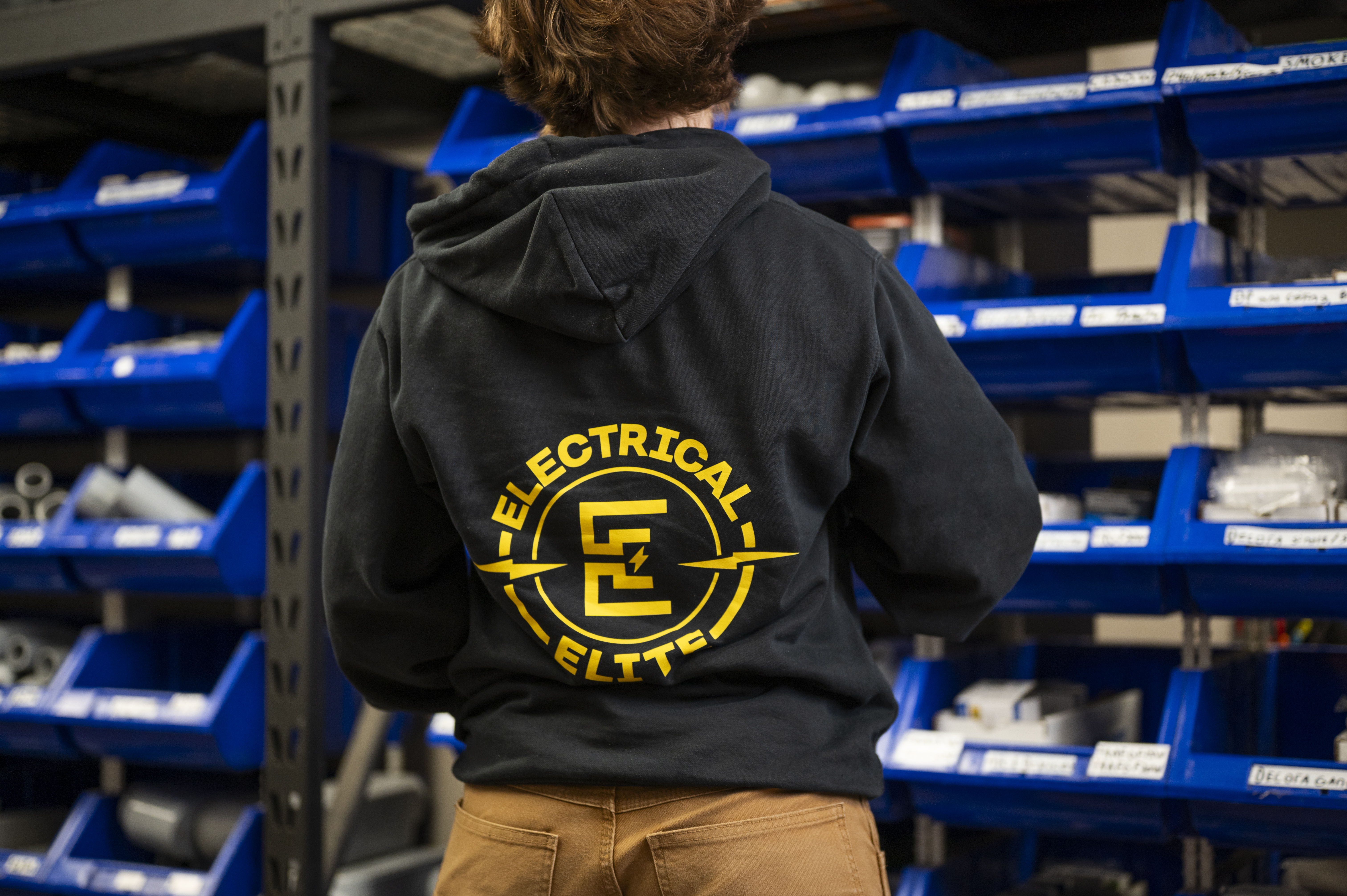 Person from behind wearing Electrical Elite hoodie in front of parts storage