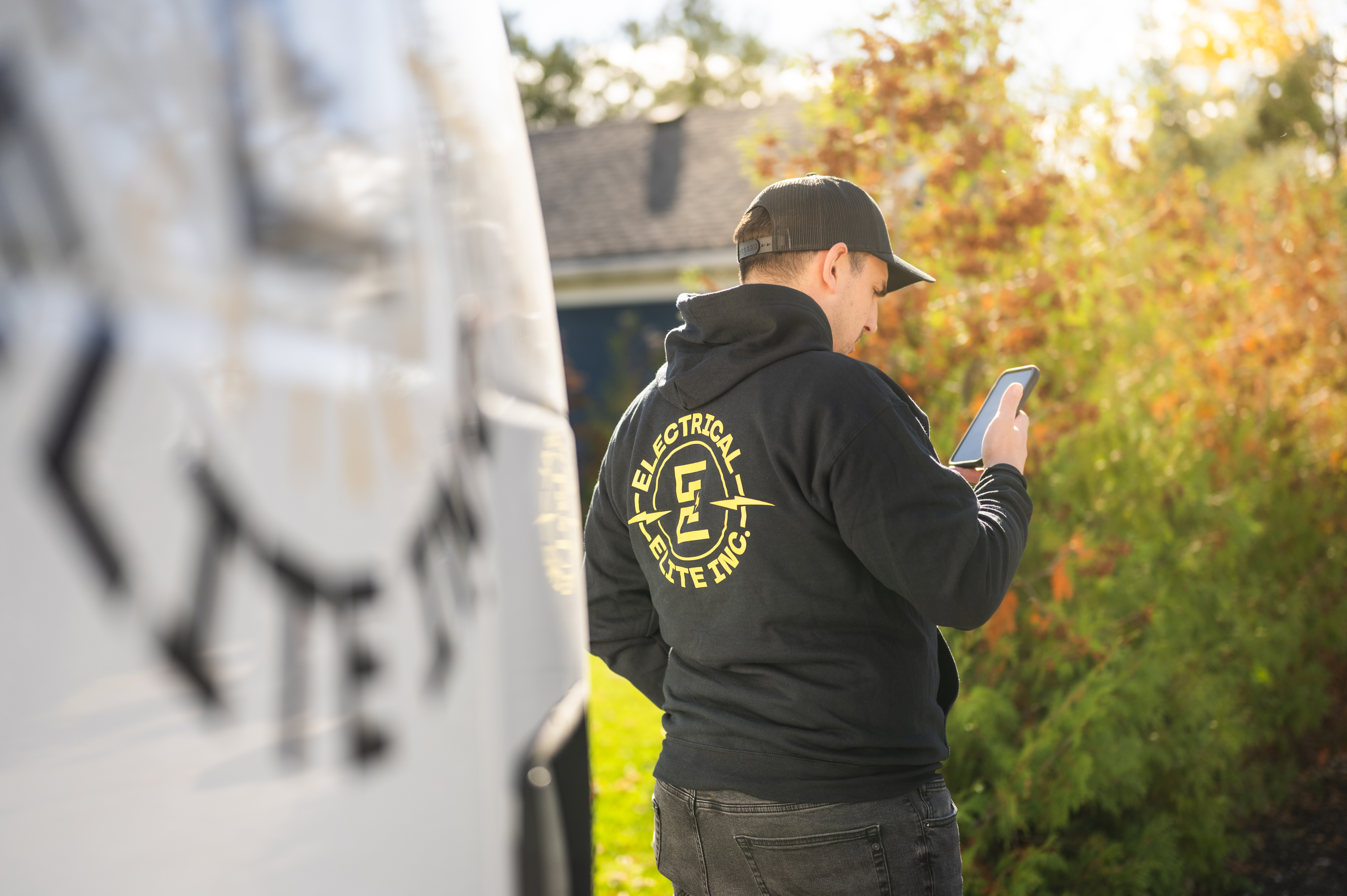 Electrical Elite employee in branded hoodie checking phone by company vehicle outdoors