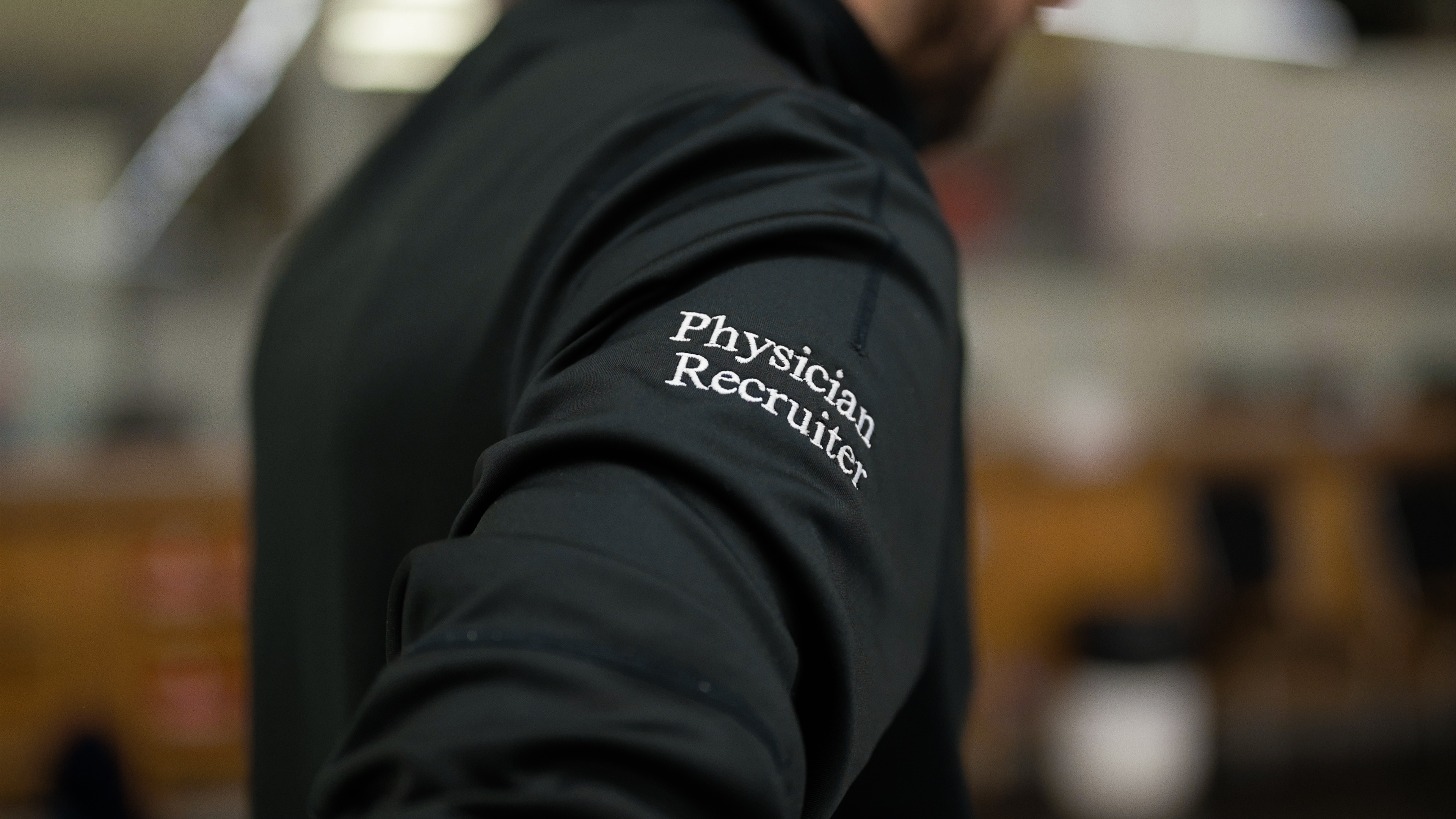 Person's arm in a black jacket with 'Physician Recruiter' embroidered on the sleeve