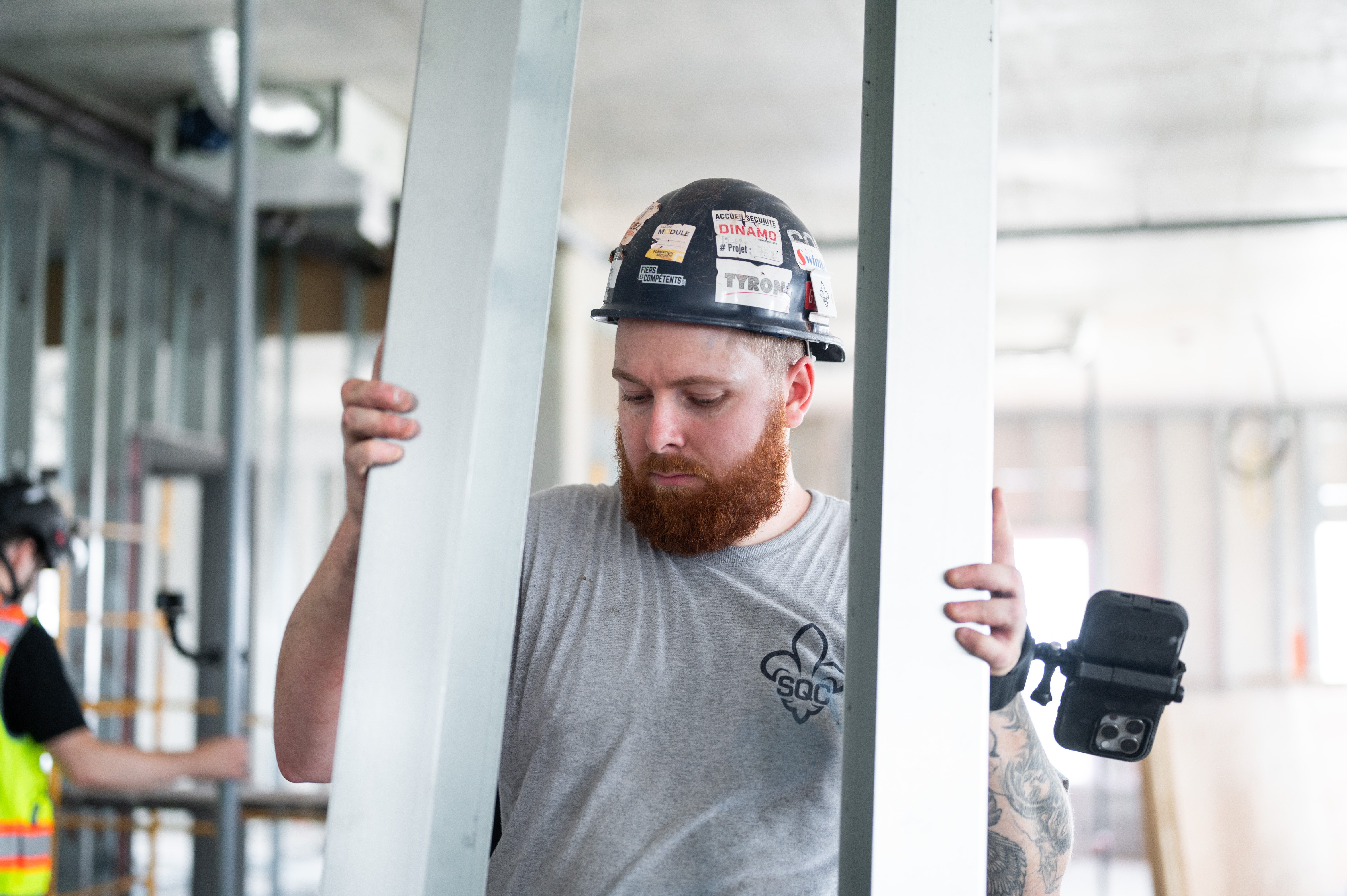 Construction worker with a red beard wearing a hard hat, carrying metal framing materials