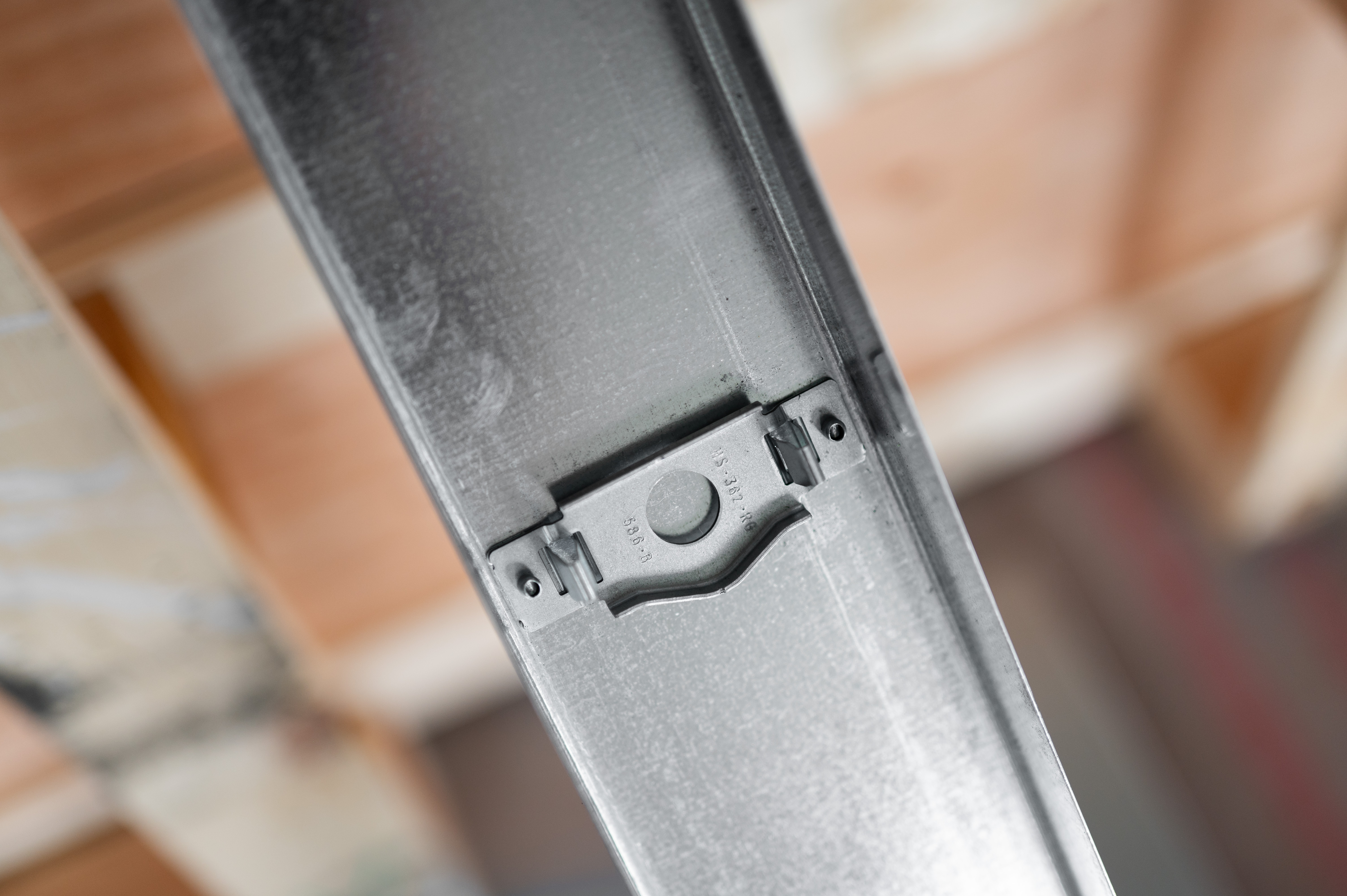 Close-up of a galvanized metal stud with a mounted bracket, against blurred wooden framing