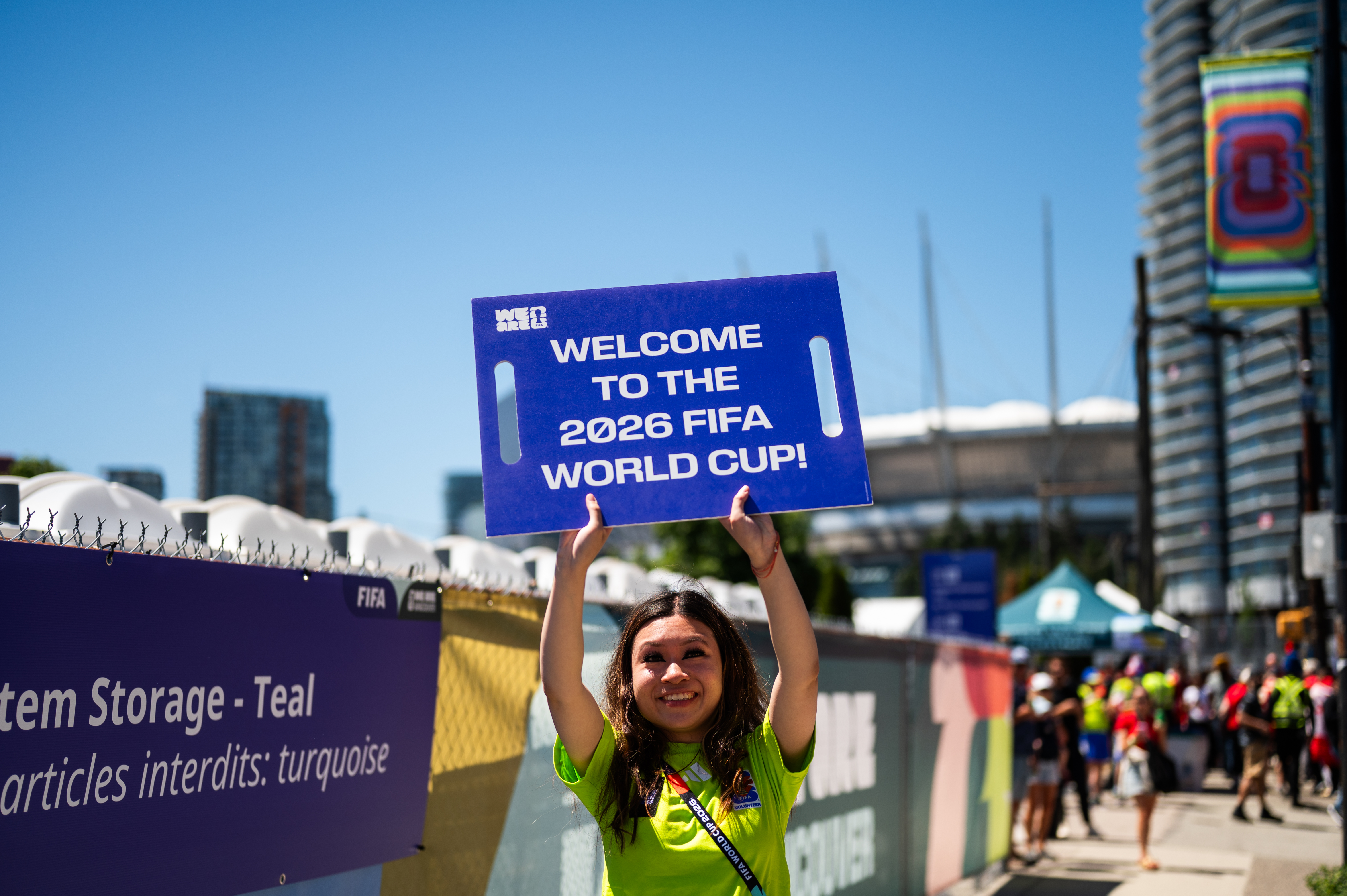 Smiling volunteer in green shirt holds 'Welcome to 2026 FIFA World Cup' sign outdoors