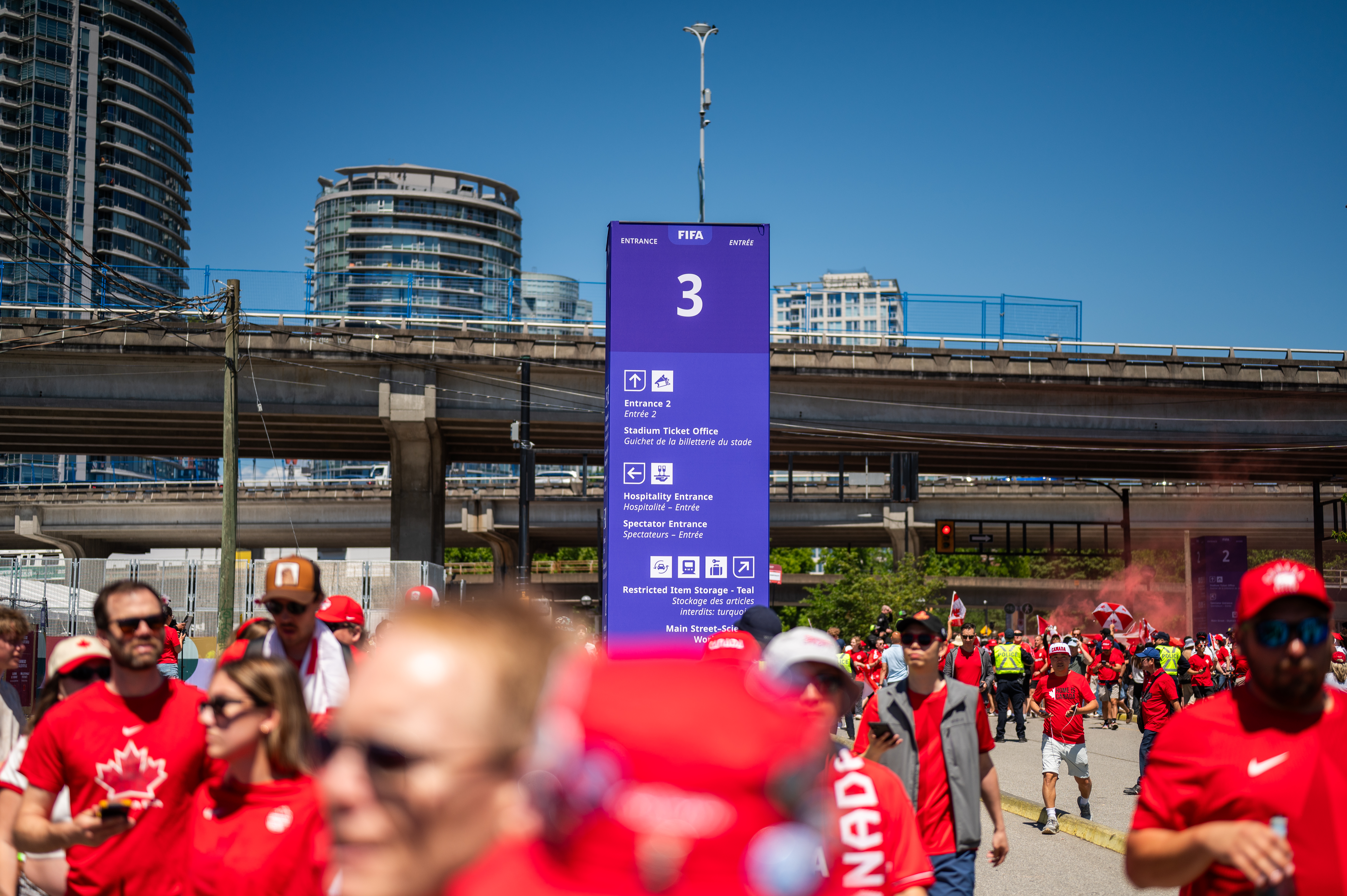 FIFA stadium directional sign 3, fans in red shirts, city buildings, World Cup event