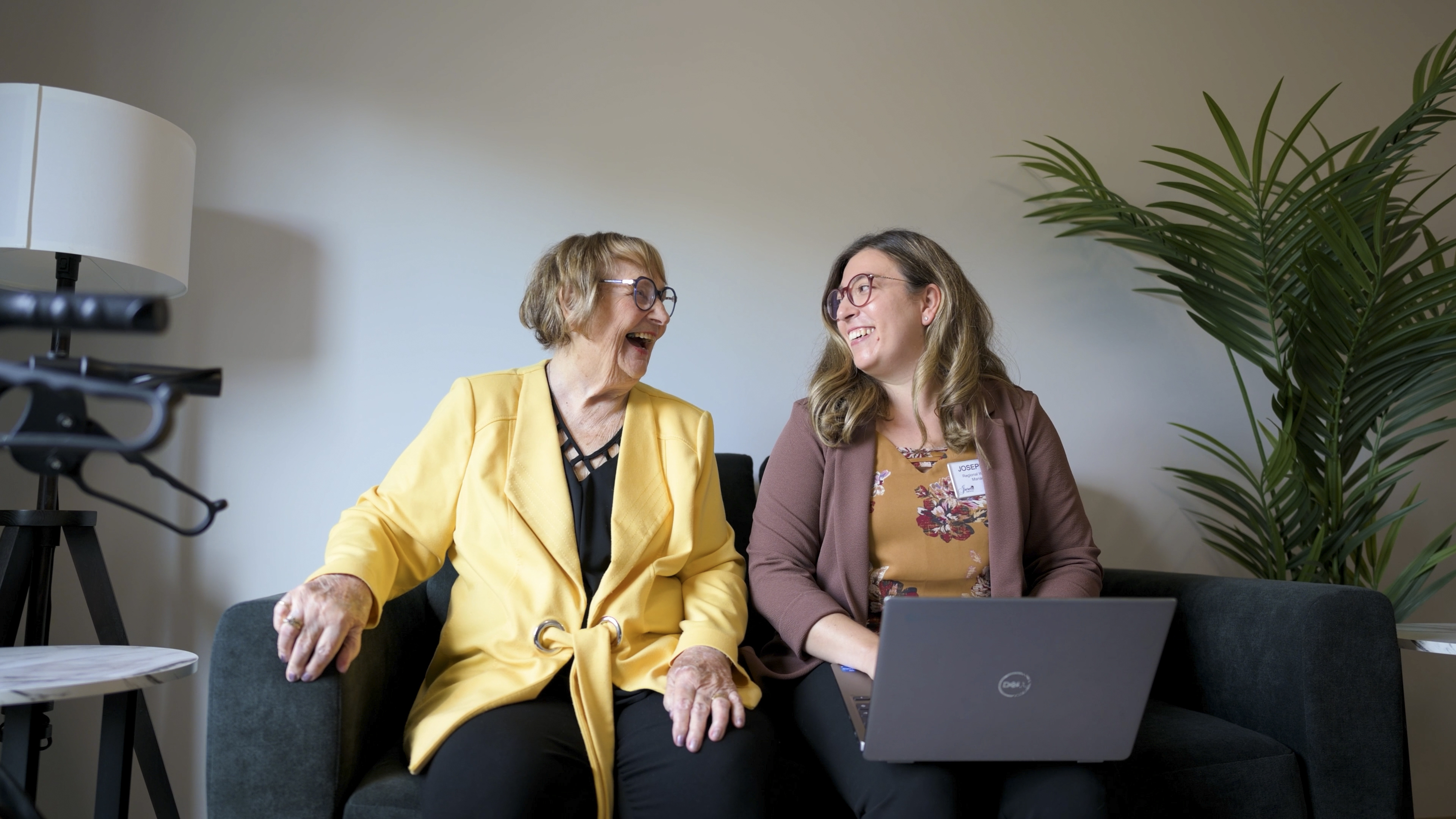Two smiling women, one older, one younger, laughing on a couch with laptop