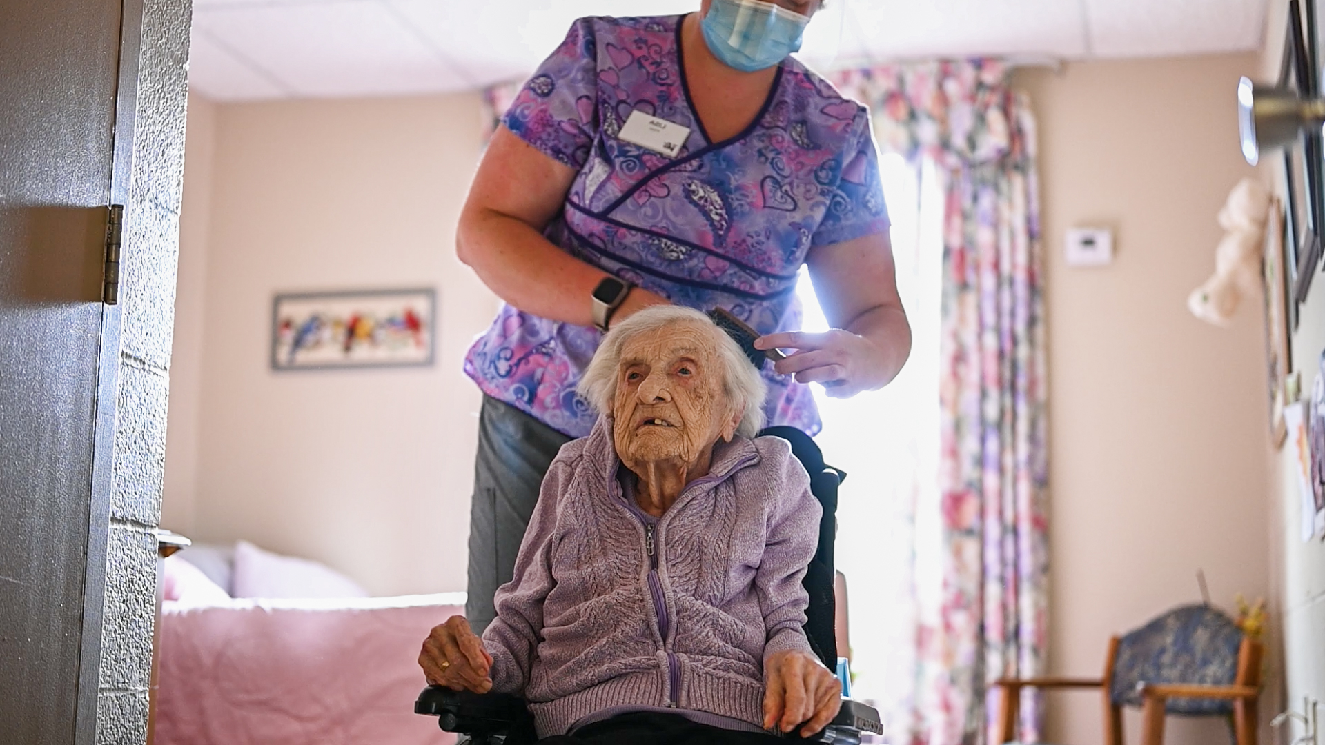Healthcare professional in scrubs brushing an elderly woman's hair in a care home